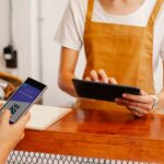 Crop anonymous female employee with application on cellphone screen interacting with partner using tablet at counter in cafeteria